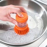 A persoa usa cepillo con mango laranxa e sedas para limpar no lavabo xabón.