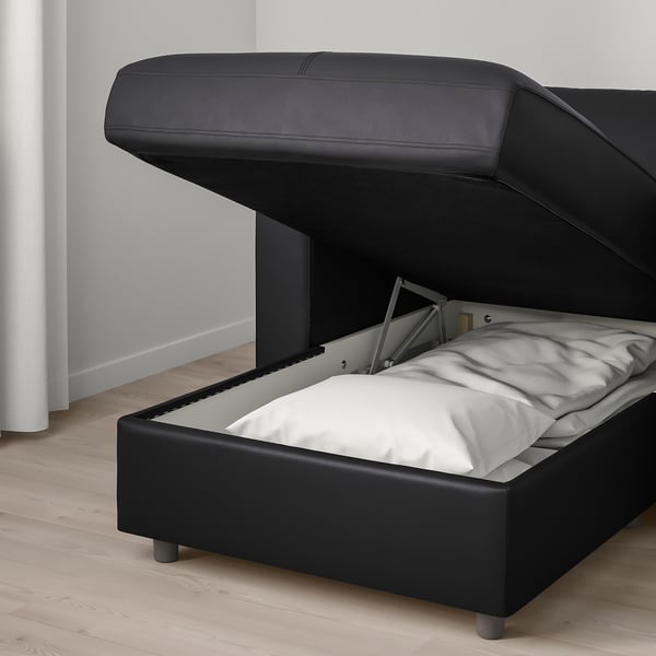 Cama de almacenamento de coiro negro con soportes metálicos, tapa aberta que revela roupa de cama branca, colocada no chan de madeira contra unha parede branca.