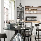 Mesa de comedor negra con cadeiras, bloc de notas, bolígrafo, vasos, cubertas VIMPELFISK, taboleiro de menú, estación de café.