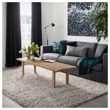 Sala de estar moderna con sofá gris, mesa de café de madeira e gran alfombra branca. Decorado con almofadas verdes e azuis e un vaso con flores brancas.