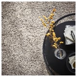 Unha alfombra VINDUM en gris claro con manchas negras. Está sentado nunha mesa escura con obxectos decorativos. A alfombra aparece suave e acolledora.