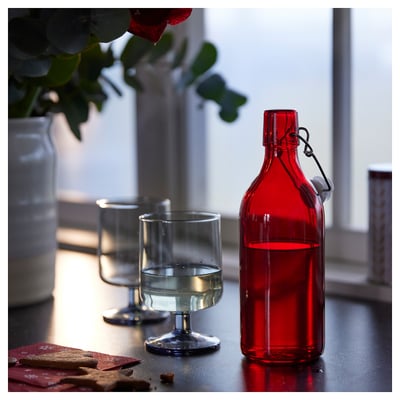Botella de vidro vermello con tapón xunto a vidro transparente nunha mesa.