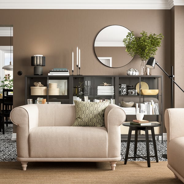 Sala de estar moderna con cadeira de brazos VISKAFORS, beige, mesa de café redonda, estante negro, lámpada de chan e alfombra beige.