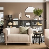 Sala de estar moderna con cadeira de brazos VISKAFORS, beige, mesa de café redonda, estante negro, lámpada de chan e alfombra beige.