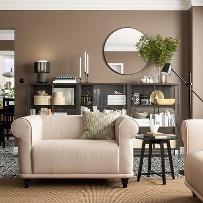 Sala de estar acolledora con cadeira de brazos VISKAFORS beige, estante negro, espello redondo e planta verde.