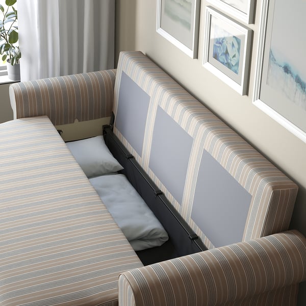 Un sofá-cama a raias móstrase parcialmente convertido nunha cama plana, revelando compartimentos de almacenamento debaixo con almofadas dentro.