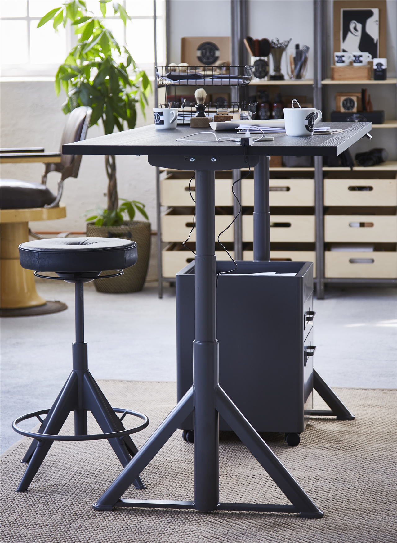Adjustable stool by a sit-and-stand desk, with diverse items on it, in an office space with a potted plant.