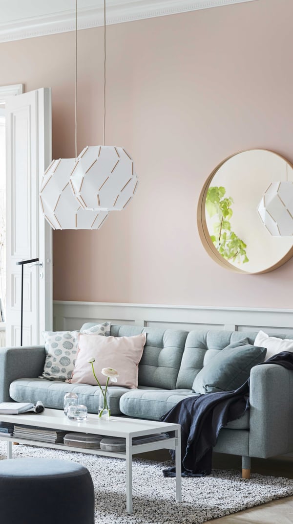 A light pink colour living room with a mirror on the wall and lamps are hanging from the ceiling. One light green sofa with pillows, throw and a white coffee table with storage in front of the sofa.