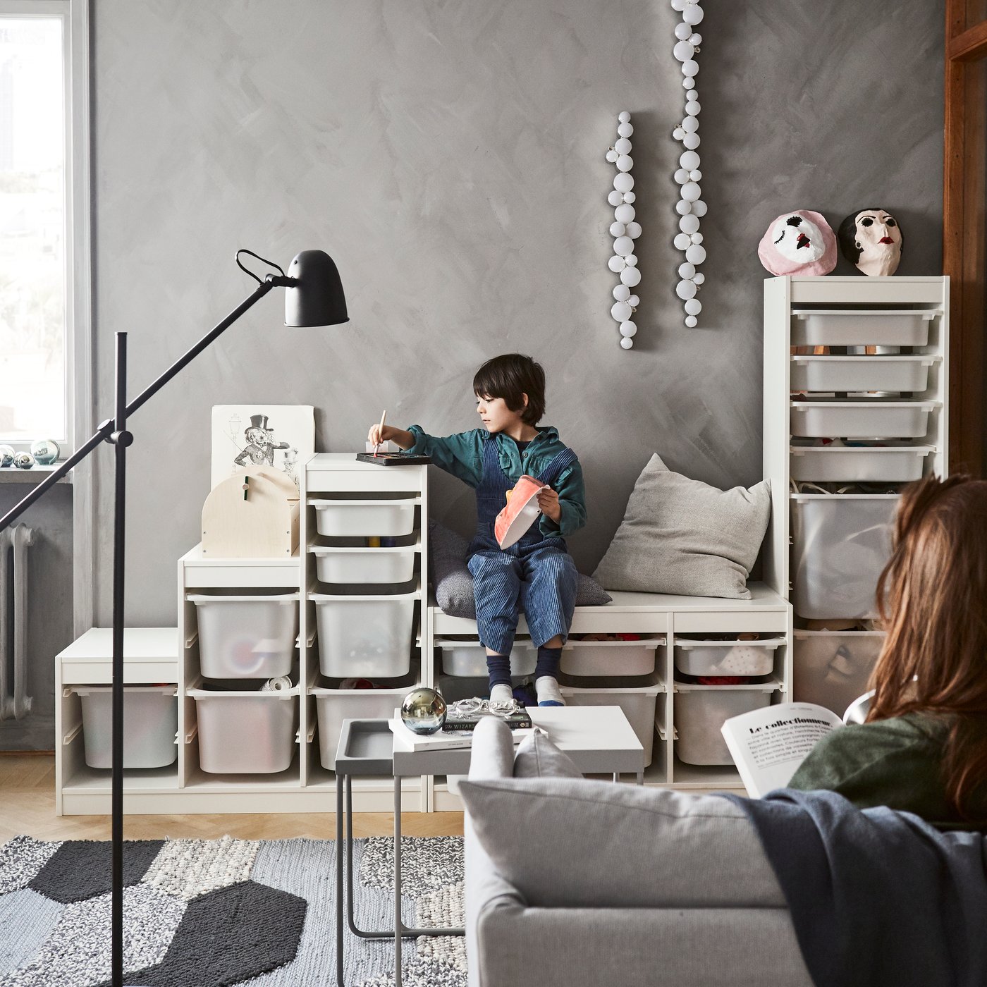 A child playing on a TROFAST combination that fills the wall as his mother sits on the sofa reading in the living room. 