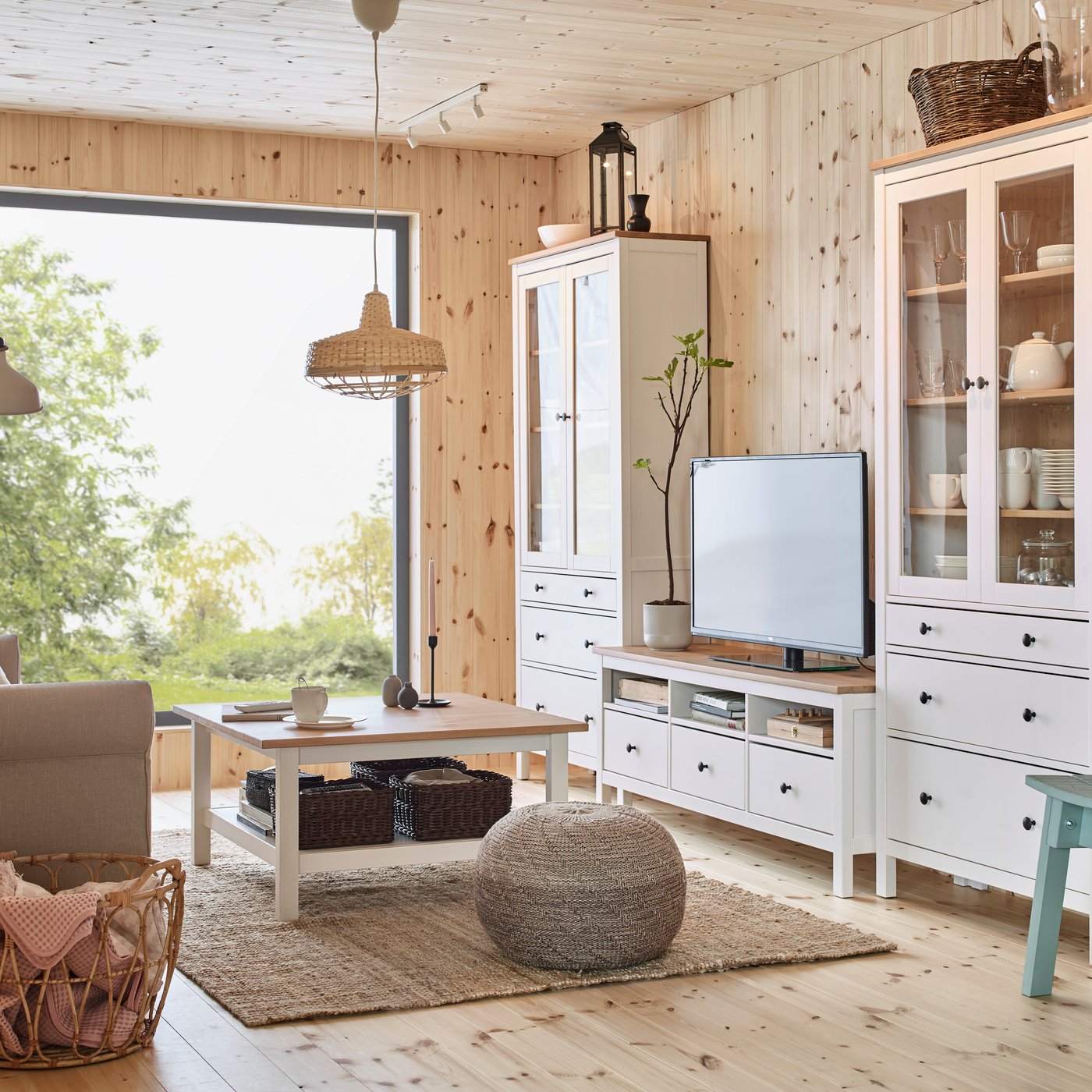 Soggiorno in stile cottage con mobili bianchi HEMNES coordinati – vetrina alta, mobile TV e madia – su pareti e soffitto in legno chiaro, grande finestra con vista sul verde, tappeto in fibra naturale, tavolino basso e dettagli in rattan e legno colorati da una sedia azzurra.