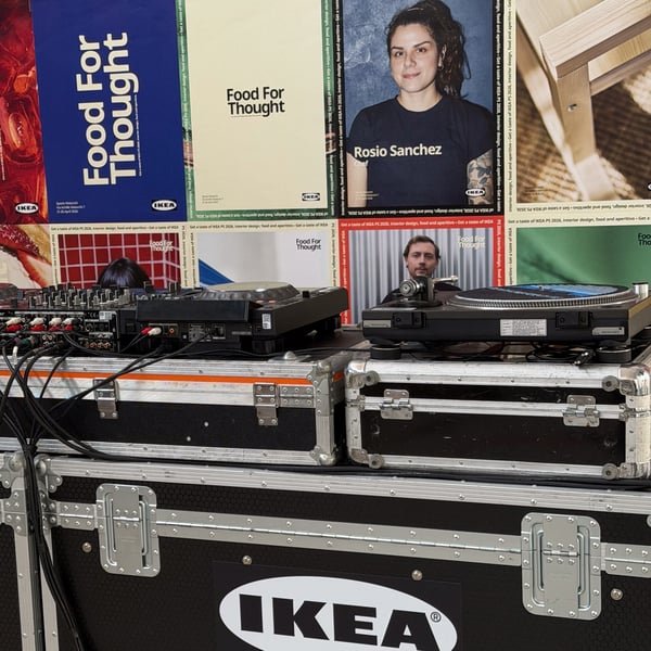 Banchi da DJ con flight case neri marchiati IKEA, mixer e piatti, davanti a una parete coperta di poster colorati.