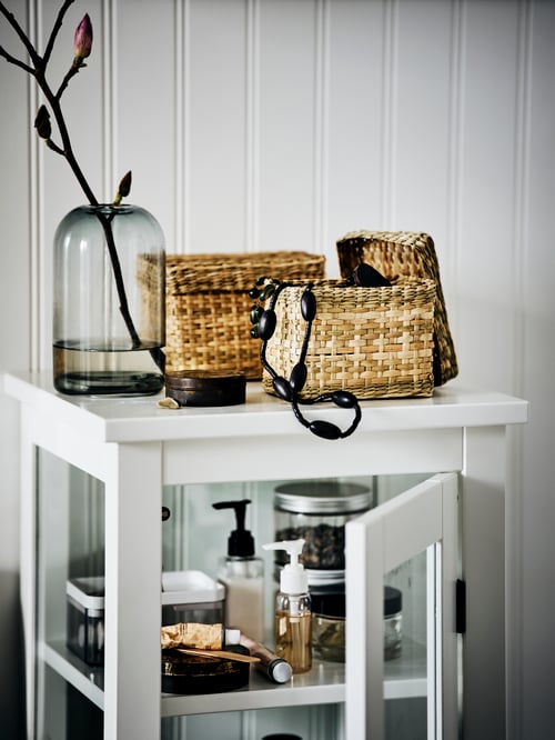 Two seagrass LURPASSA boxes by a vase on top of a HEMNES high cabinet. One box is open with a necklace hanging out.