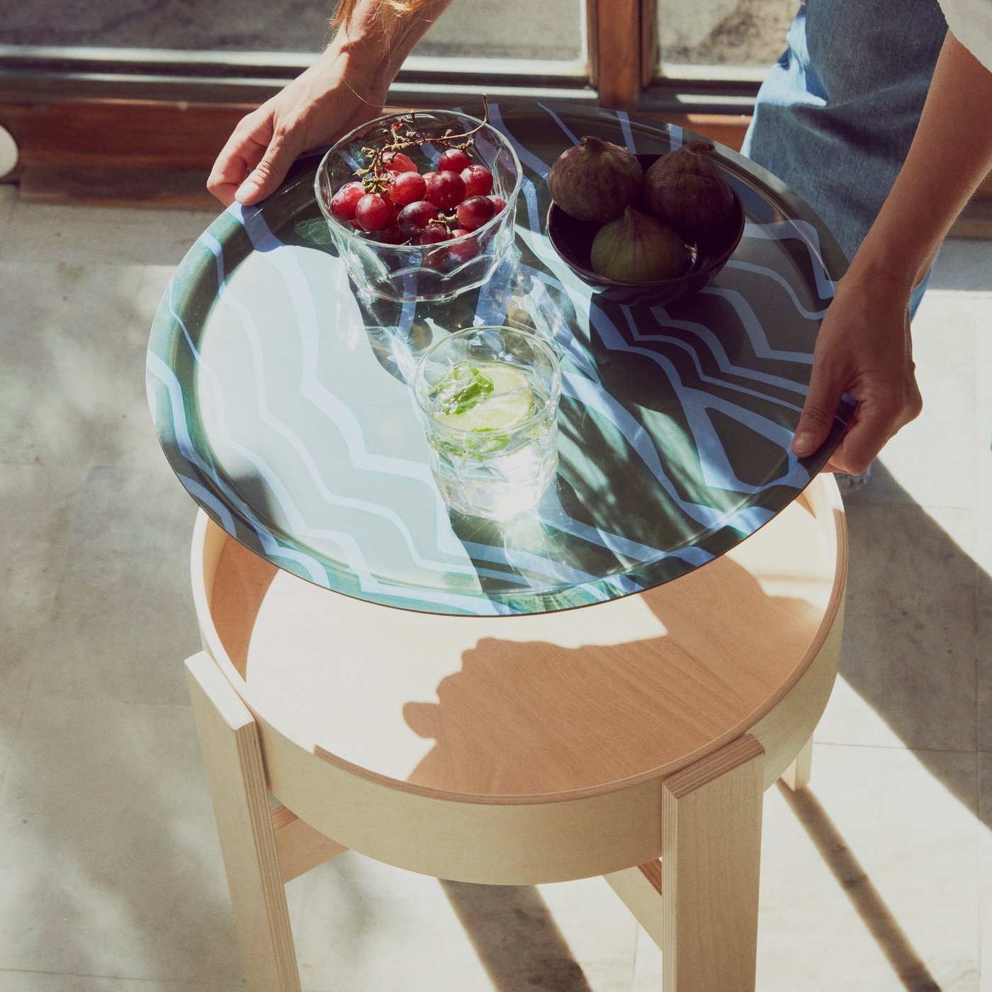 Una persona che sta posando su un tavolino BASTUA un vassoio multicolore a fantasia con bevande e stuzzichini.