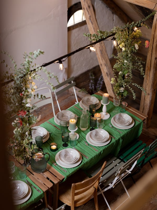 A cozy, rustic dining table set with green linen, glassware, candles, and floral decorations in a wooden-beamed room.