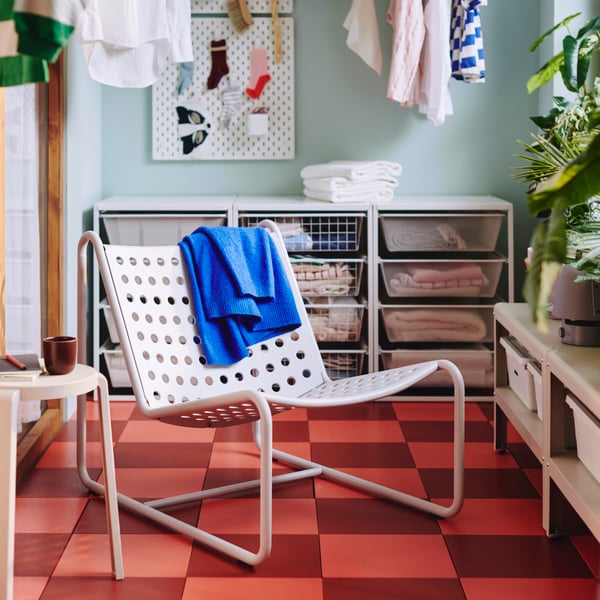 An enclosed balcony has a light grey coated-steel, outdoor SKOGSÖN easy chair with a perforated seat, near low open storage.