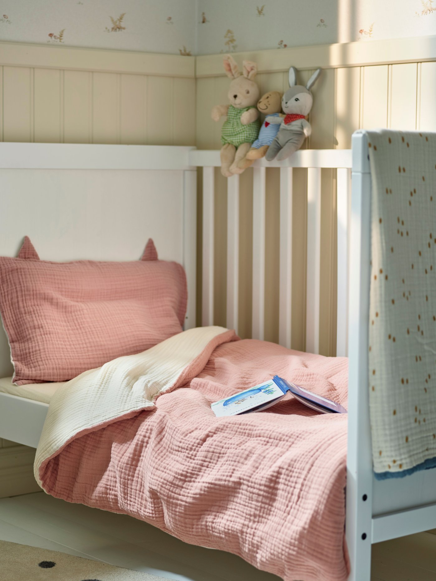 Una cuna blanca con ropa de cama rosada y una manta GULDVÄVARE.