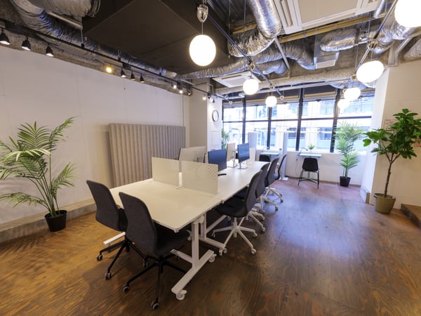 白いデスクと黒い椅子が並ぶ明るいオフィス。木の床と観葉植物が温かみを添える協働スペース