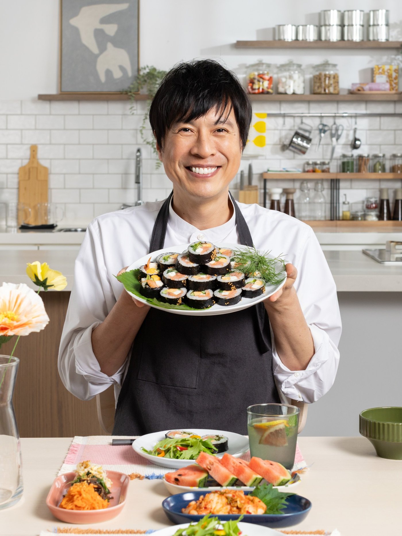 家庭のキッチンでエプロンを着た人が、巻き寿司やサラダなど彩り豊かな寿司レシピの料理をテーブルに並べて持っている。