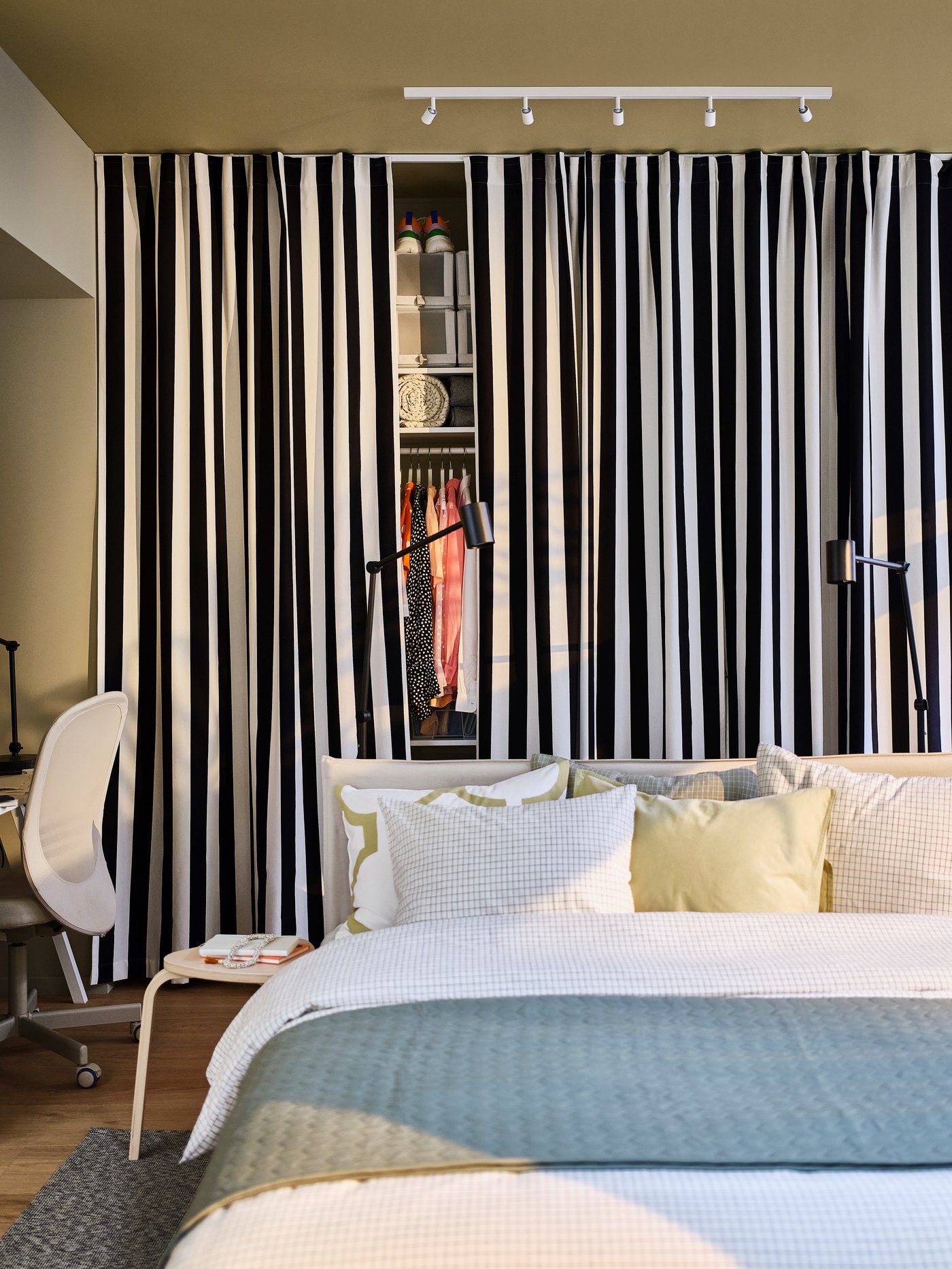 Sovrum med bäddad säng i ljusa textilier, svartvita randiga gardiner som döljer en garderob med kläder, samt skrivbordsstol och läslampor vid sängen.
