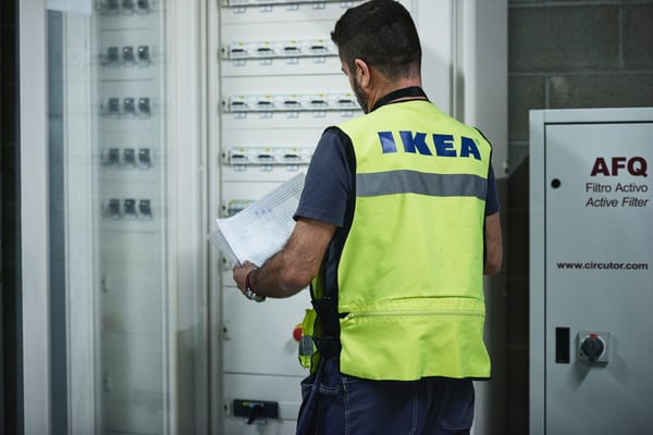 An IKEA co-worker wearing a yellow high-visibility vest is looking at drawings in a large electrical cabinet.