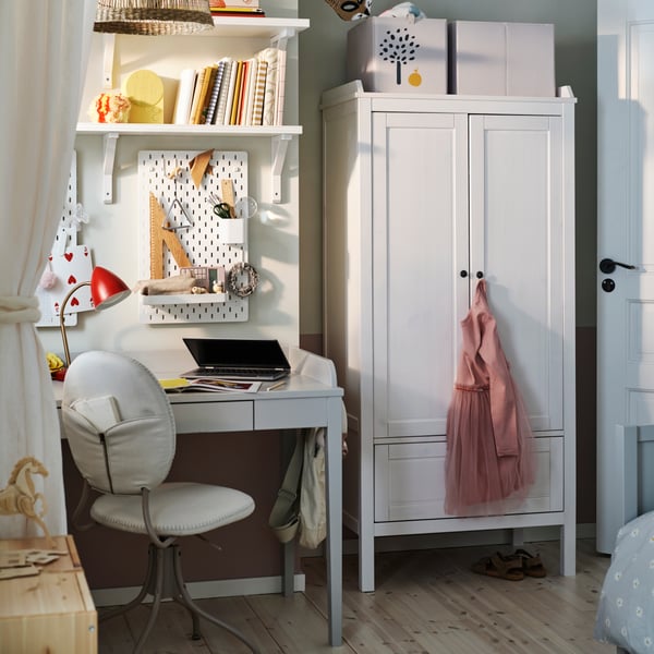 Ein weisser SUNDVIK Kleiderschrank steht in einem Kinderzimmer neben einem grauen SMYGA Schreibtisch mit einem Regal darüber und einem BJÖRKBERGET Stuhl.
