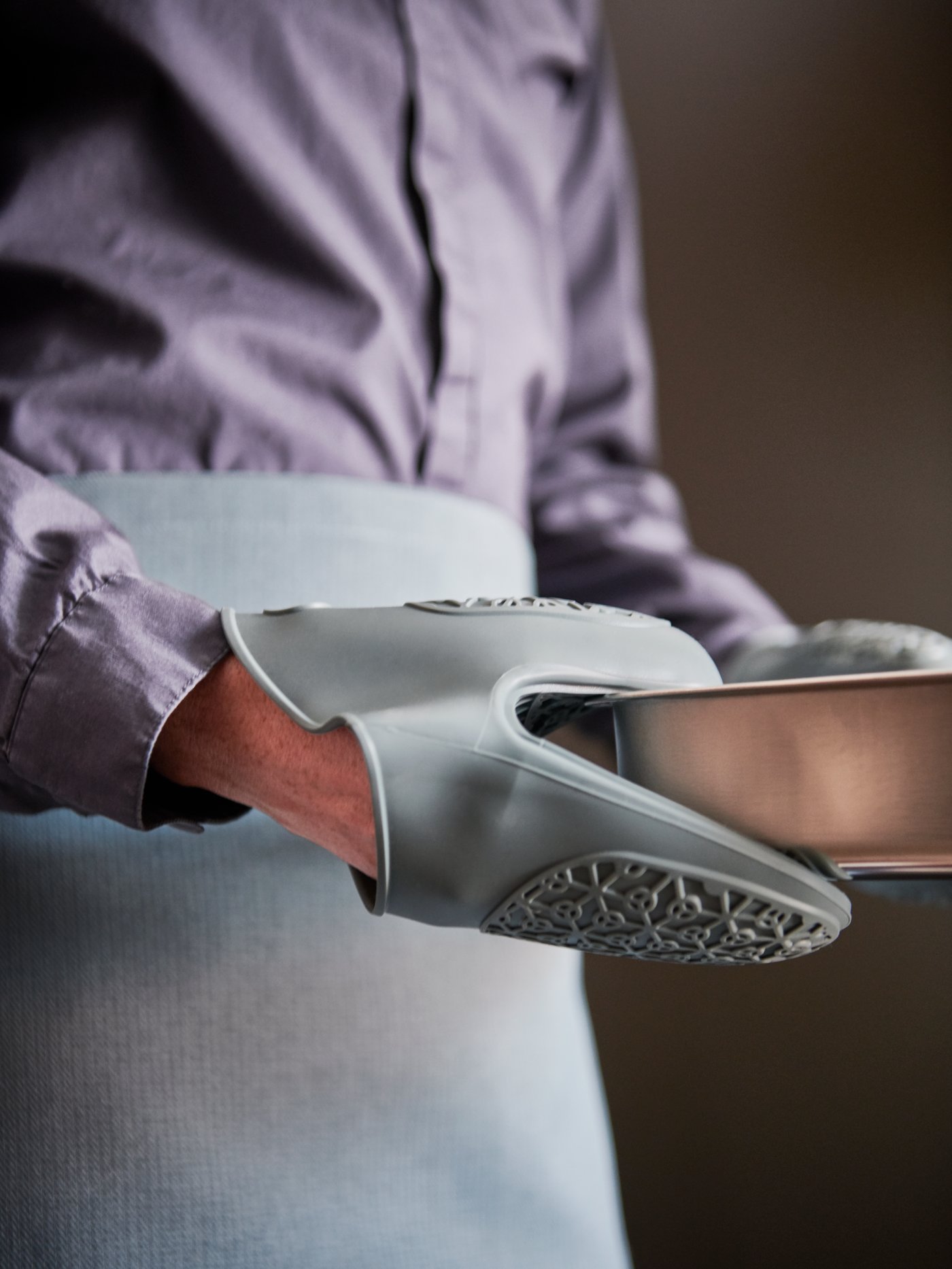 A person with a blue SANDVIVA waist apron holding a grey HEMMABAK roasting tin with a blue SANDVIVA silicone oven glove.