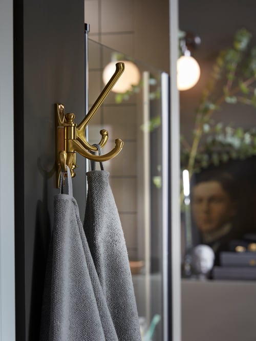 Une salle de bains avec un crochet mobile triple couleur laiton placé à côté d’une cabine de douche, avec deux serviettes de bain grises accrochées.