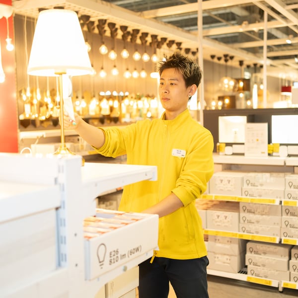 照明売場で、黄色いユニフォームのイケアのコワーカーが点灯したランプを指しながら商品説明をしている
