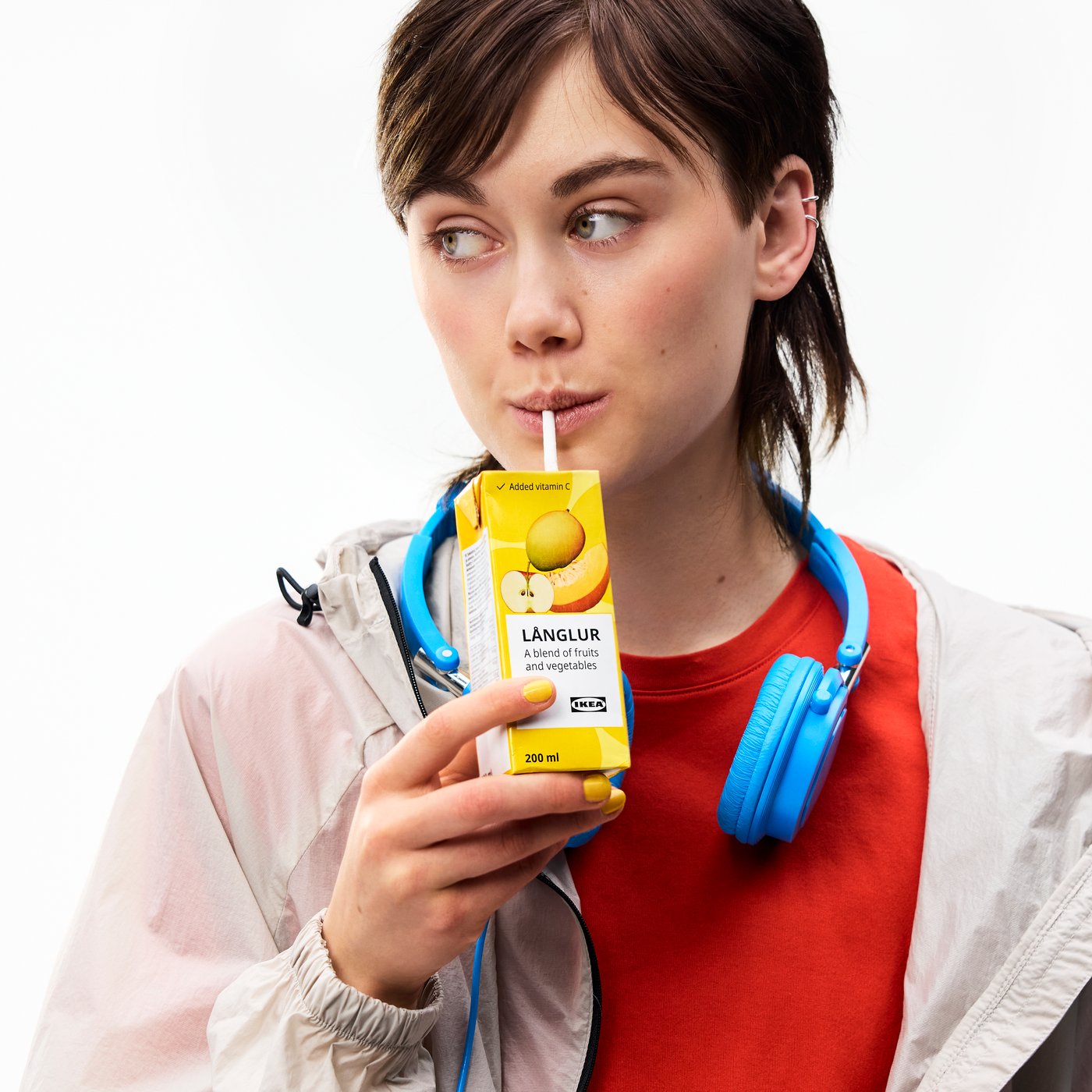 A teenager is sipping a LÅNGLUR fruit and vegetable smoothie from a straw, with a pair of headphones around their neck.