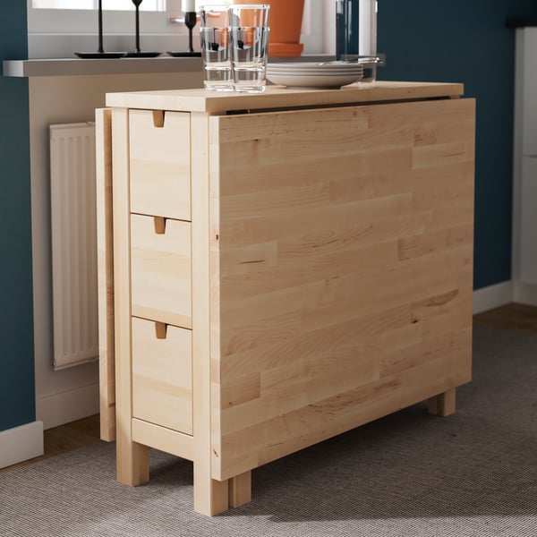 A birch NORDEN gateleg table with its surface folded is standing against a wall, with plates and glasses on it.