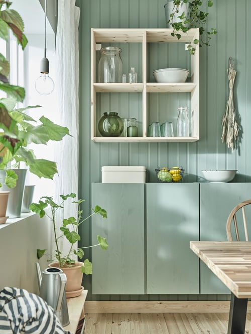An IVAR shelving unit is mounted on a green wall. Under the unit is an IVAR cabinet with green doors. Plants are to the side.