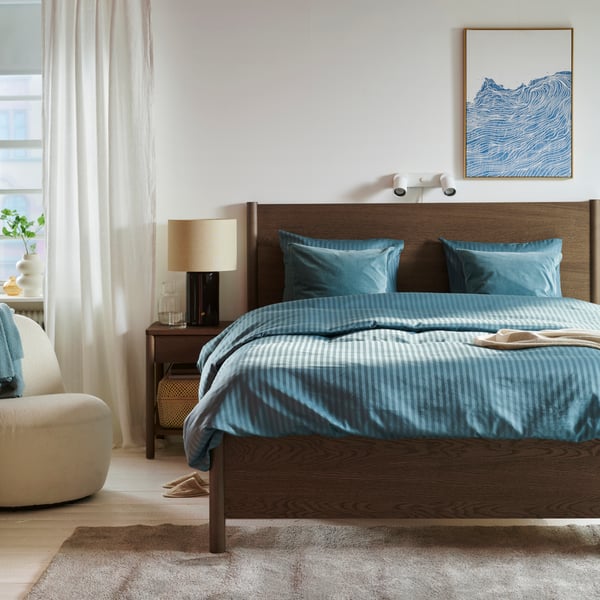 A bright bedroom with white walls and a dark brown TONSTAD bed with a blue NATTJASMIN duvet cover.