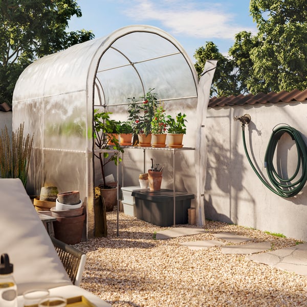 A transparent white ASPAREN greenhouse with rounded edges is in a sunny garden. Inside are herb-filled terracotta pots.