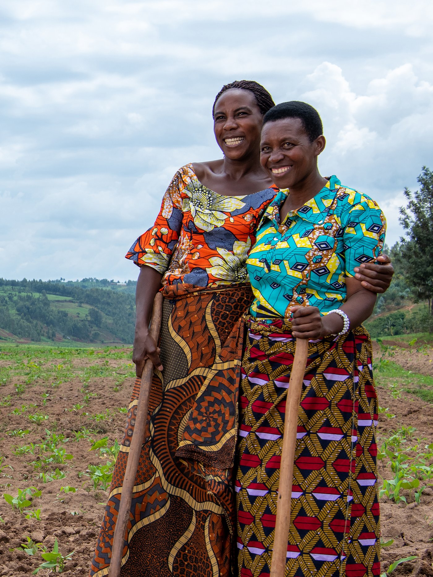 Two people stand in a field with arms around each other, holding rakes and wearing colourful clothes.