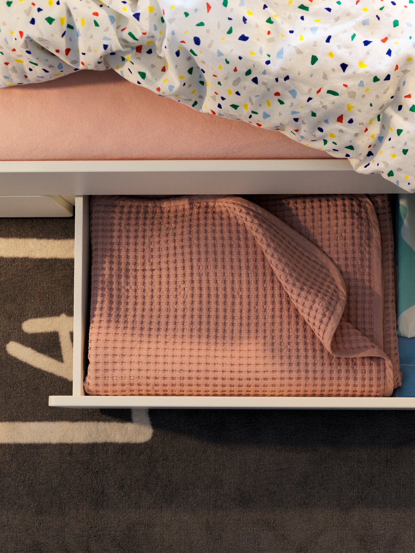 Detalle de un dormitorio infantil con una estructura de cama blanca, un cajón de almacenaje, una colcha, una funda nórdica y una funda de almohada con un estampado y una alfombra. 