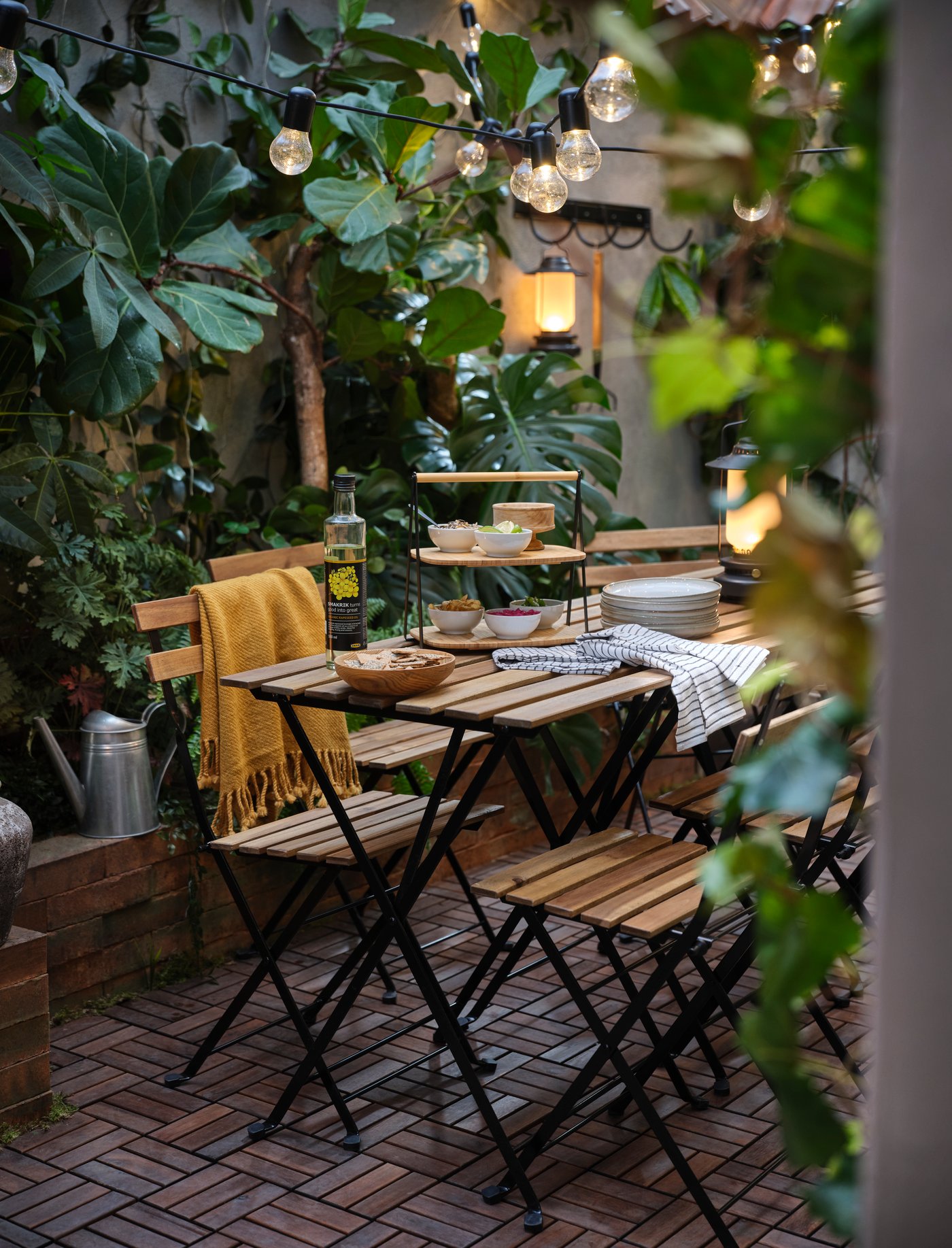 Uteplats med träbord, stolar på tegelstensyta, dukat med porslin och serveringsfat. Omgiven av gröna växter och hängande ljusslingor.