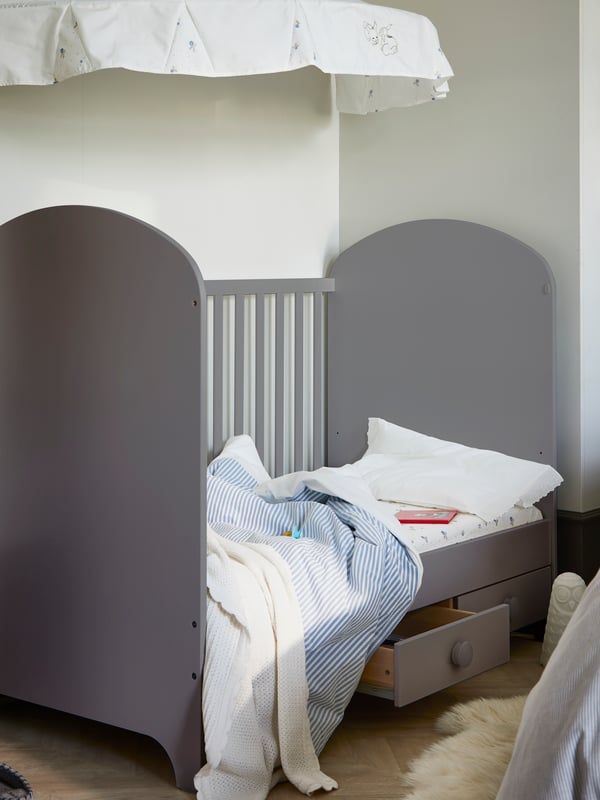 A light grey GONATT cot that has one side removed and has one drawer pulled out stands in a corner under a RÖDHAKE canopy.