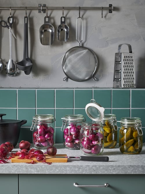 Eine Küchenarbeitsplatte mit KORKEN Dosen mit Deckeln, in denen Gurken und rote Zwiebeln eingemacht sind. An der Wand darüber hängt eine Reihe von Utensilien.