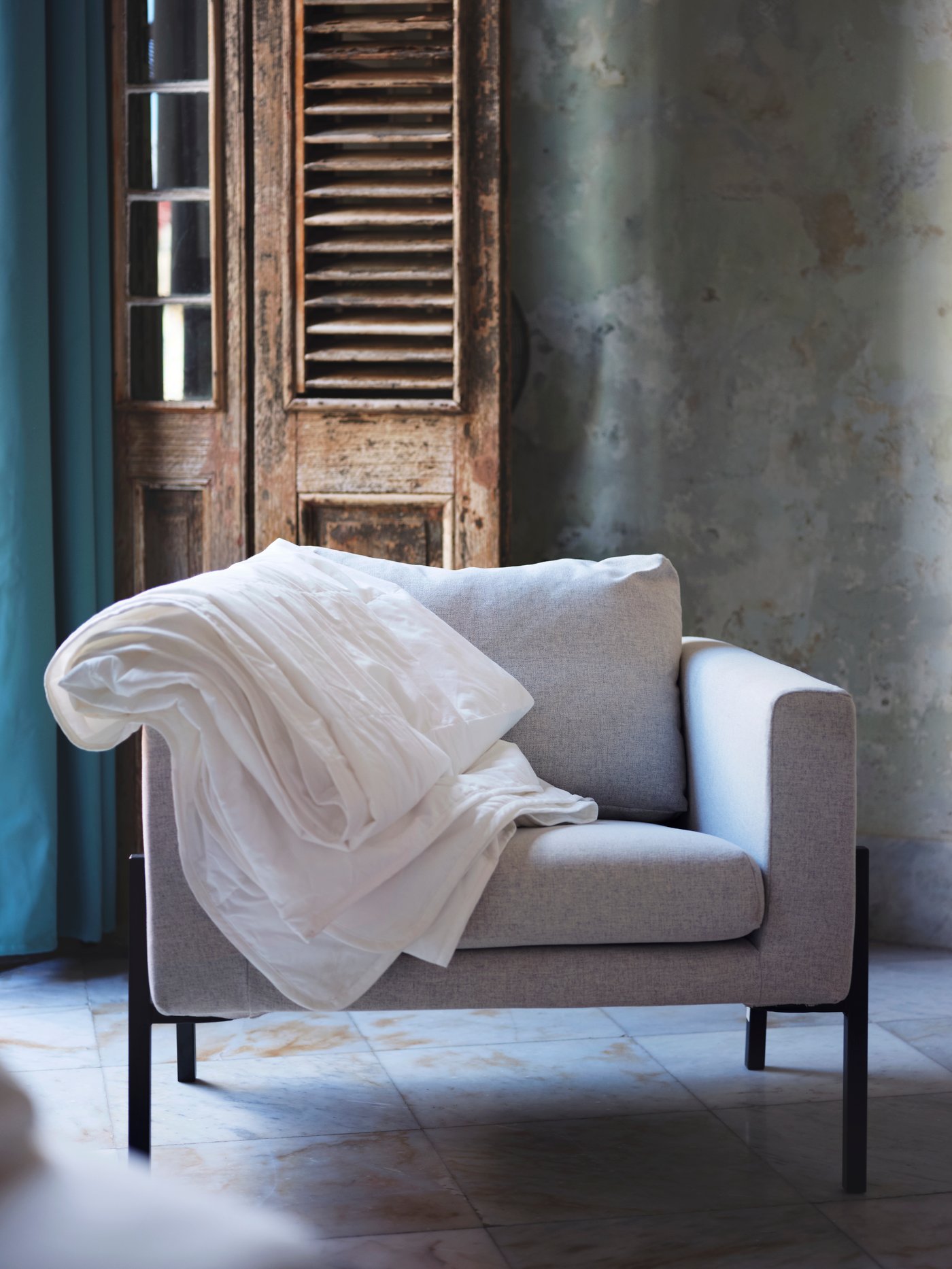 A grey chair with a white duvet draped over one arm, set against a rustic, wooden door on a tiled surface.