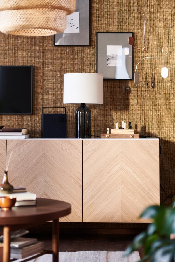 An oak veneer BESTÅ cabinet in a living room by a brown wall with picture, a TONVIS lamp and Bluetooth a speaker on top.