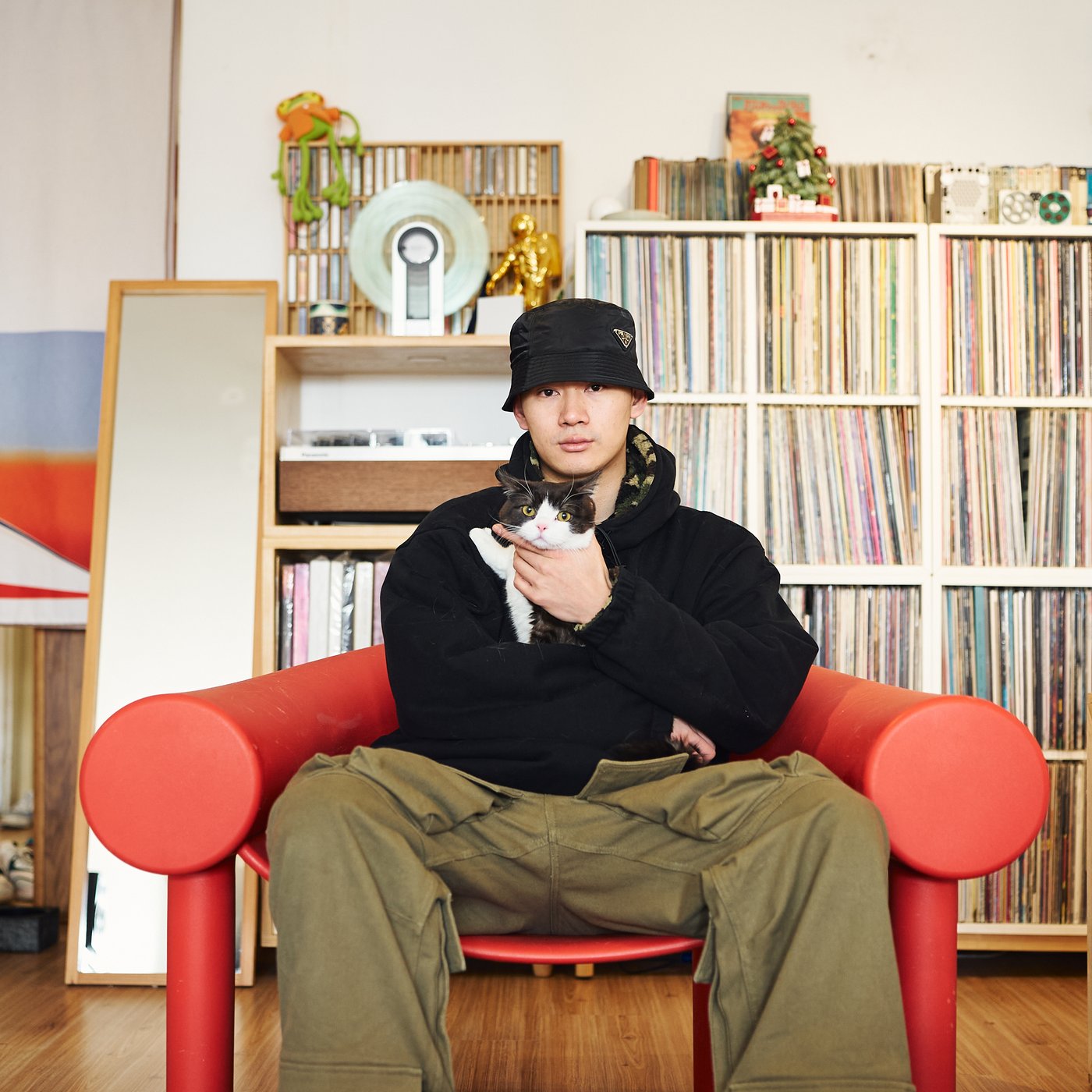A man in a hat is sitting in a red armchair with a cat in his arms. There is a big shelving unit behind him full of vinyl records.