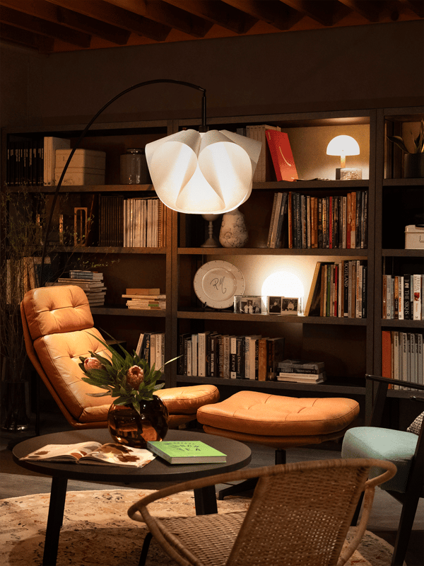 本棚と間接照明に囲まれたリビングの一角。キャメル色のラウンジチェアとオットマン、丸テーブルに花瓶と本が置かれたくつろぎスペース。