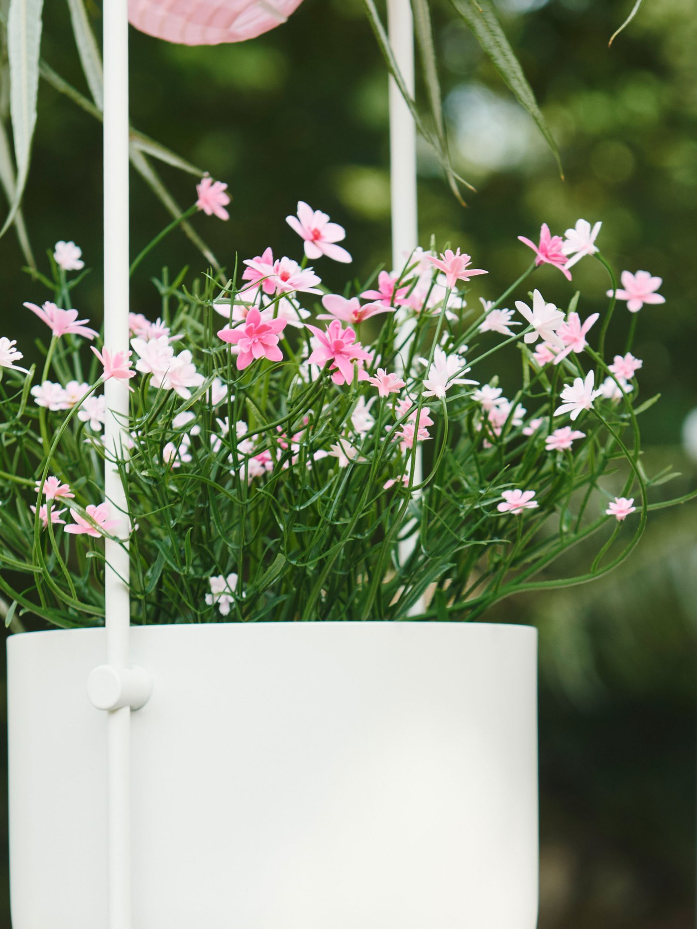 Närbild på konstgjord krukväxt med små rosa blommoe i vit ampel.