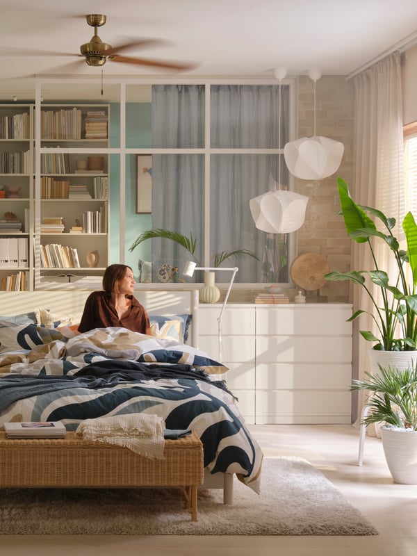 A person is sitting in a bed made with MYGGLASVINGE bed linen in a bright bedroom with chest of drawers, plants and more.