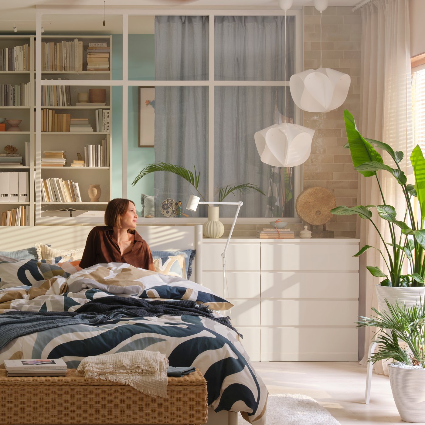 A person is sitting in a bed made with MYGGLASVINGE bed linen in a bright bedroom with chest of drawers, plants and more.