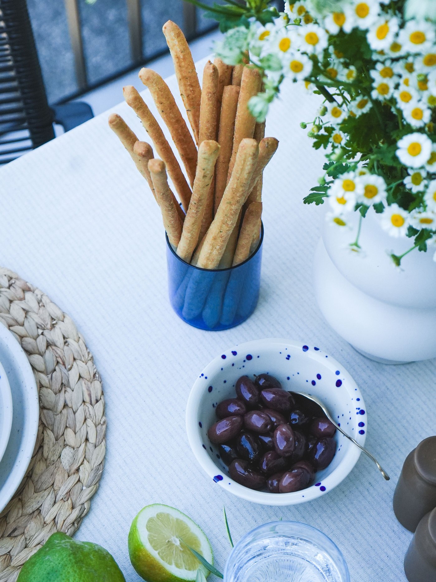 Une table dressée avec des grissini dans un verre bleu, une tasse SKUGGSTUBB ornée de pâquerettes blanches, un bol SILVERSIDA rempli d'olives noires, de citrons frais et de citrons verts.