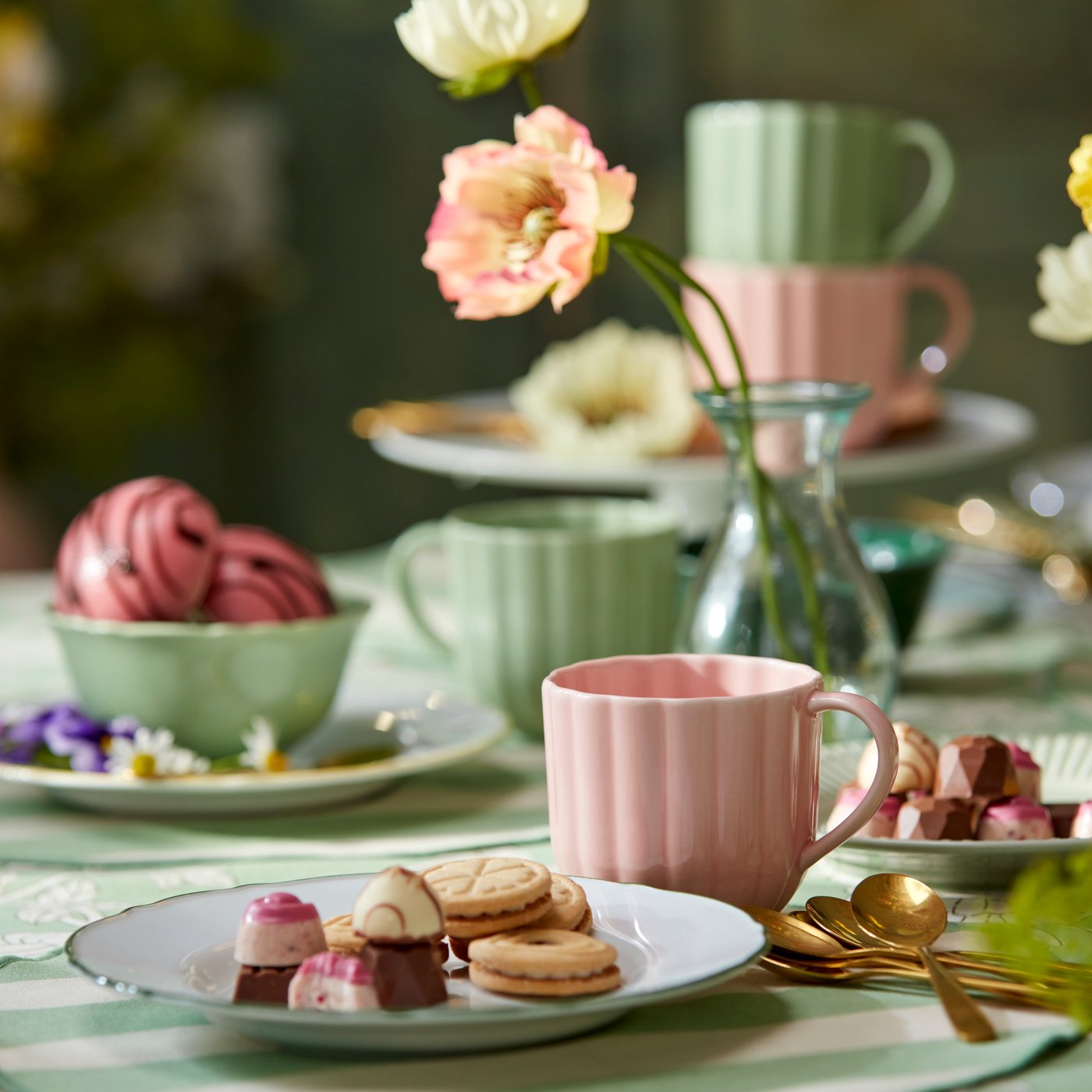 Pastellfärgad dukning med rosa kopp, tallrik med praliner och småkakor, gröna koppar i bakgrunden och blommor i glasvas på bordet.