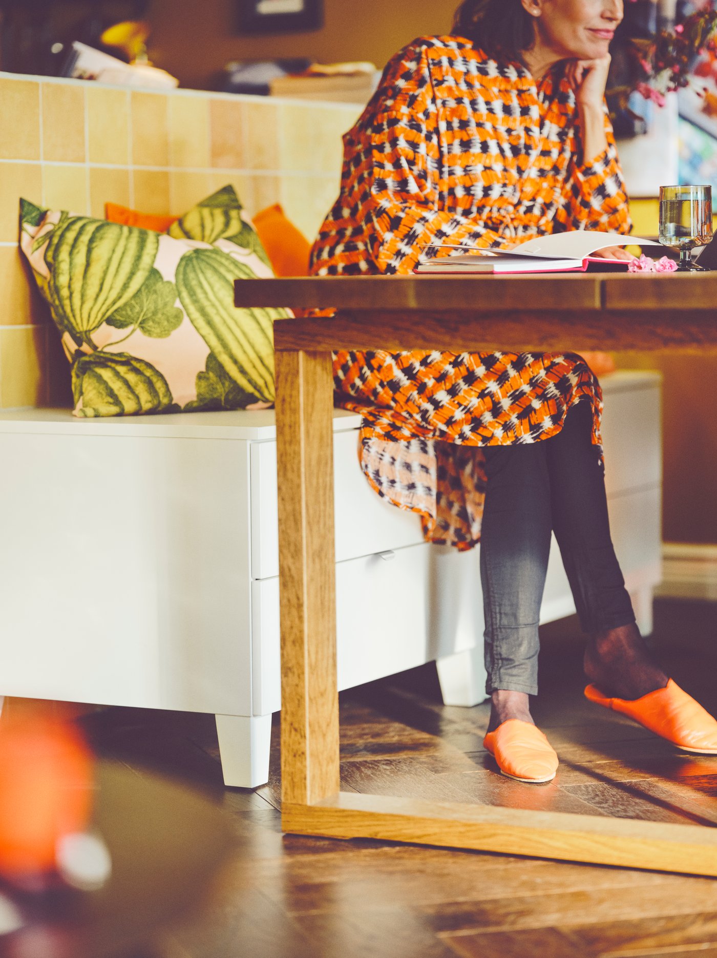 Une personne est assise à une table MÖRBYLÅNGA en bois, sur un rangement PLATSA blanc garni de coussins. Un livre et un verre sont posés sur la table.