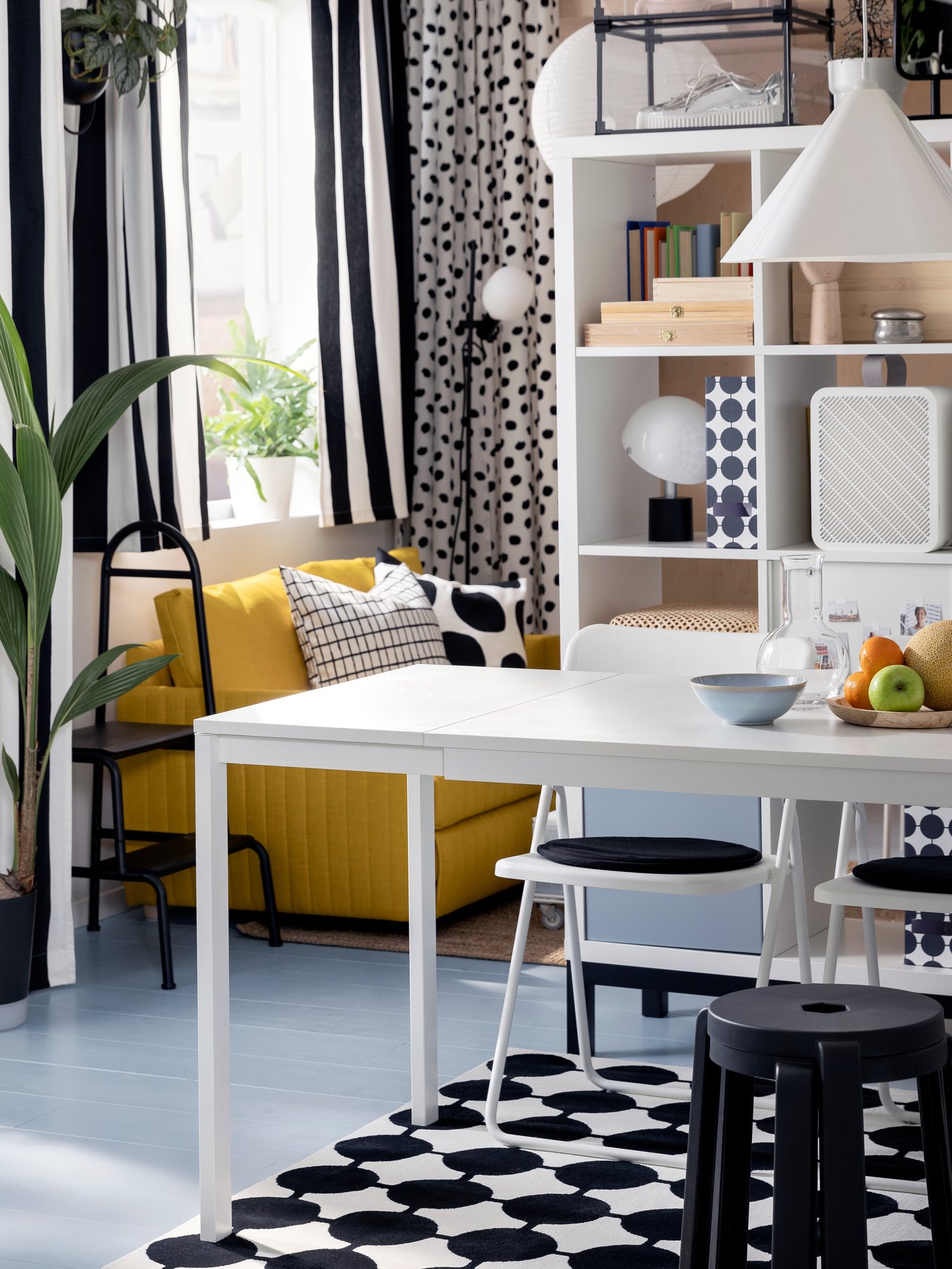 Två staplade, mörkgrå IBBARBRO pallar står vid ett vitt utdragbart VIHALS bord framför en vit KALLAX hyllenhet.