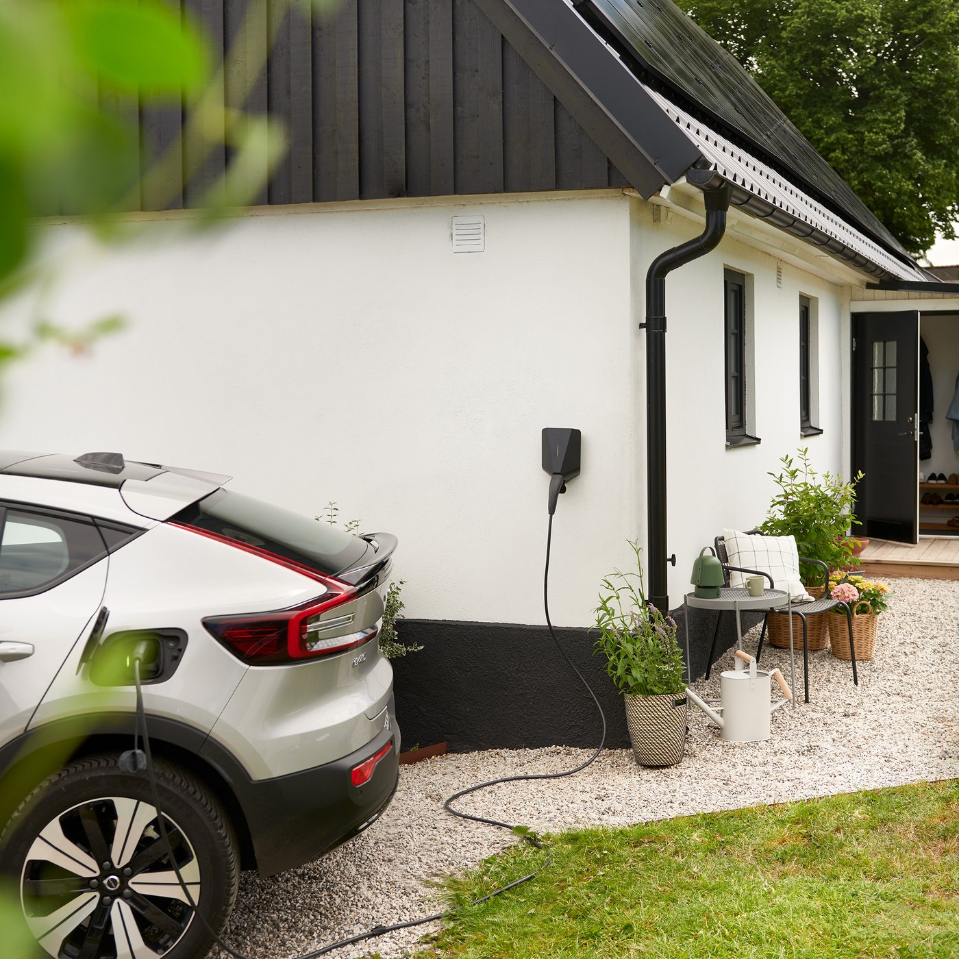 Een moderne woning met witte muren en een zwart dak, waar een elektrische auto in de oprit staat te laden aan een laadstation naast het huis.
